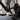 A close-up of a black indoor cycling trainer mounted on a bicycle frame, with a pedal, chainring, and flywheel visible in a home gym setting. A water bottle and folded towel sit on the floor nearby.