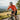 A cyclist riding on a country road while drinking from a white water bottle, wearing a bright orange short-sleeve cycling jersey, black shorts, a white helmet, and sunglasses.
