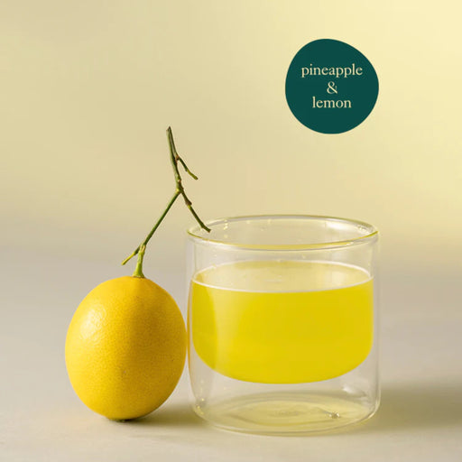 Clear glass tumbler with yellow liquid next to a pineapple on a light background