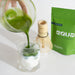 Green liquid being poured into a glass with a matcha whisk and packaging in the background.