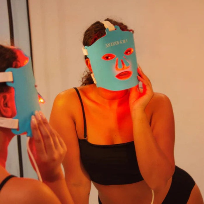 Person wearing a blue LED face mask in front of a mirror with red light.
