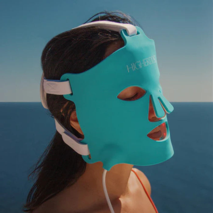 Person wearing a blue face mask with 'HIGHER' branding against a blue sky and ocean background