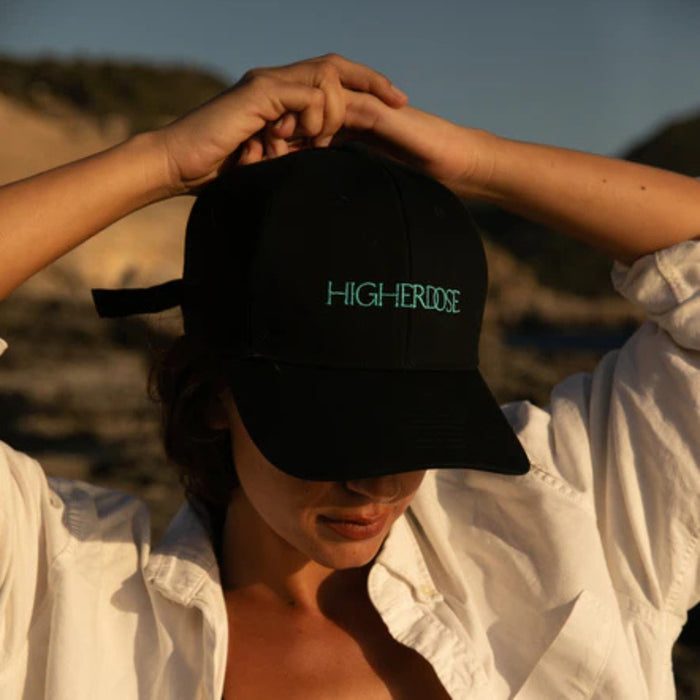 Person wearing a black cap with 'HIGHERDOSE' on it, outdoors.