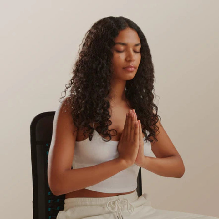 Woman meditating with hands pressed together in a chair against a plain background