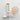 A white Precision Hydration 1000 supplement tube with orange and green text, shown on a light gray background alongside a small glass of pale amber liquid, a white cap, and a white stand.