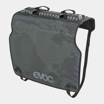 A black EVOC tailgate pad designed to secure two bicycles during transport on a pickup truck. The pad features a patterned design with the brand logo visible.